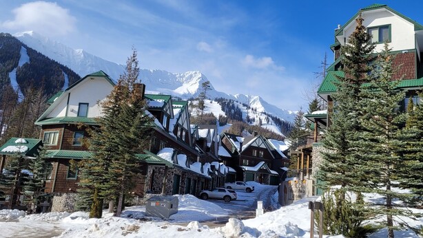 Une ville de montagne enneigée, avec des bâtiments de style chalet traditionnel, une piste de ski en arrière-plan et un ciel bleu dégagé.