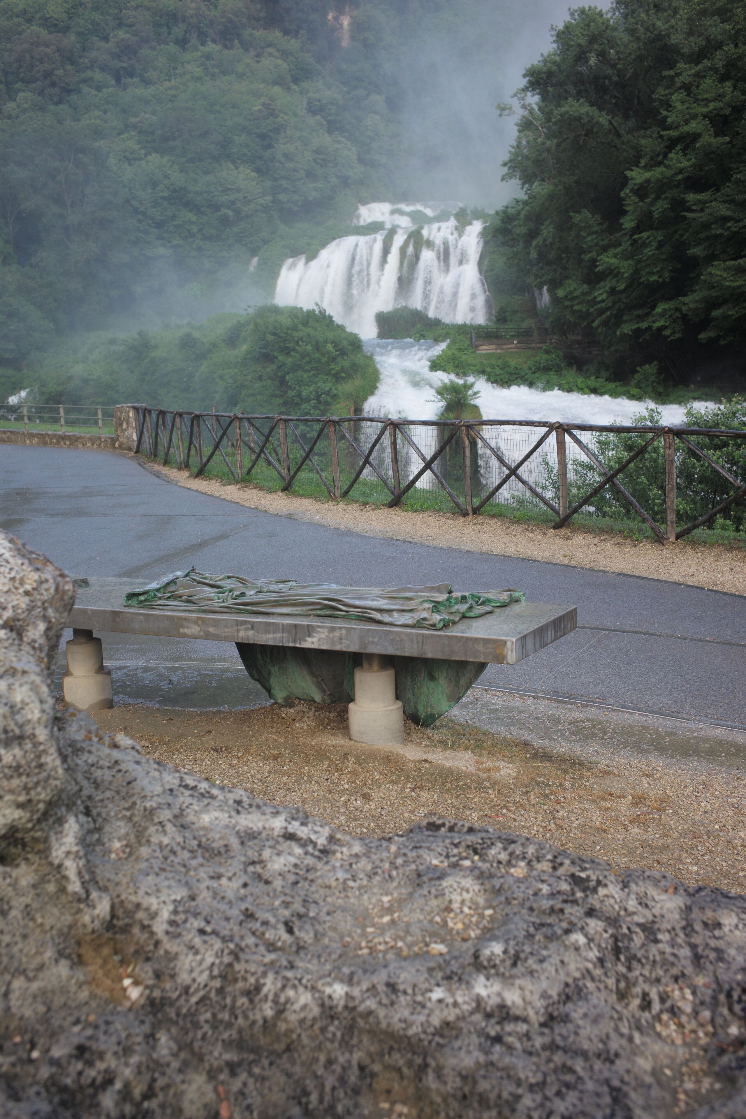 A paved road with a metal railing, a concrete bench, and a waterfall in the background.