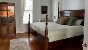 Une chambre à coucher avec un lit à baldaquin en bois, un radiateur, une armoire aux portes vitrées et un portrait accroché au mur.
