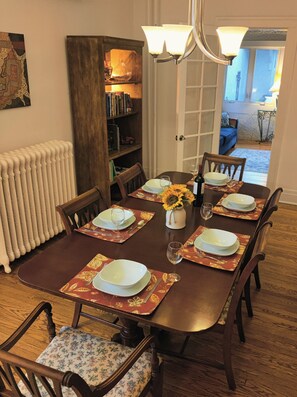 A dining room with a wooden table set for six, surrounded by chairs with floral upholstery. A bookshelf and a radiator are visible in the background.