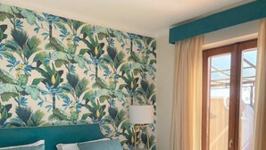 Una habitación de hotel con una cama grande, una silla transparente, una mesita redonda y un papel tapiz tropical.