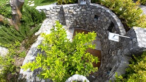 Un espacio al aire libre con paredes de piedra, un suelo de madera, rodeado de vegetación y una estructura de piedra con chimenea.