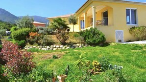 A yellow house with a covered porch and a garden with various plants and a rock border. - Villa Roccu (Borgo)