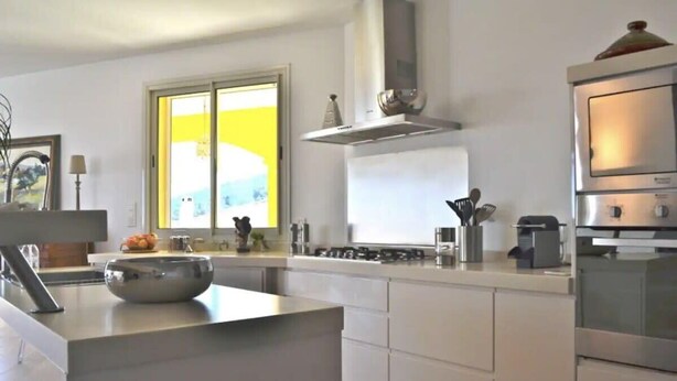 A modern kitchen with a central island, stainless steel appliances, and a large window with yellow curtains. - Villa Roccu (Borgo)
