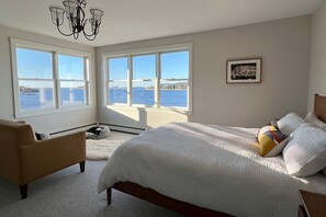 A bedroom with a large bed, two windows with a view of the sea, a chandelier, and a framed picture on the wall.