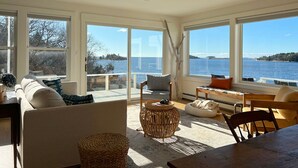 Ein geräumiges Wohnzimmer mit großem Fenster und Blick auf ein Gewässer, ausgestattet mit einer Couch, Sesseln und einem kleinen Tisch.