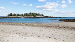 Ein Strand mit Kieselsteinen und eine felsige Insel mit Bäumen im Hintergrund.