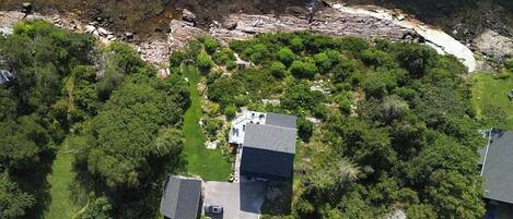 An aerial view of a residential area with houses surrounded by dense greenery and a rocky shoreline.