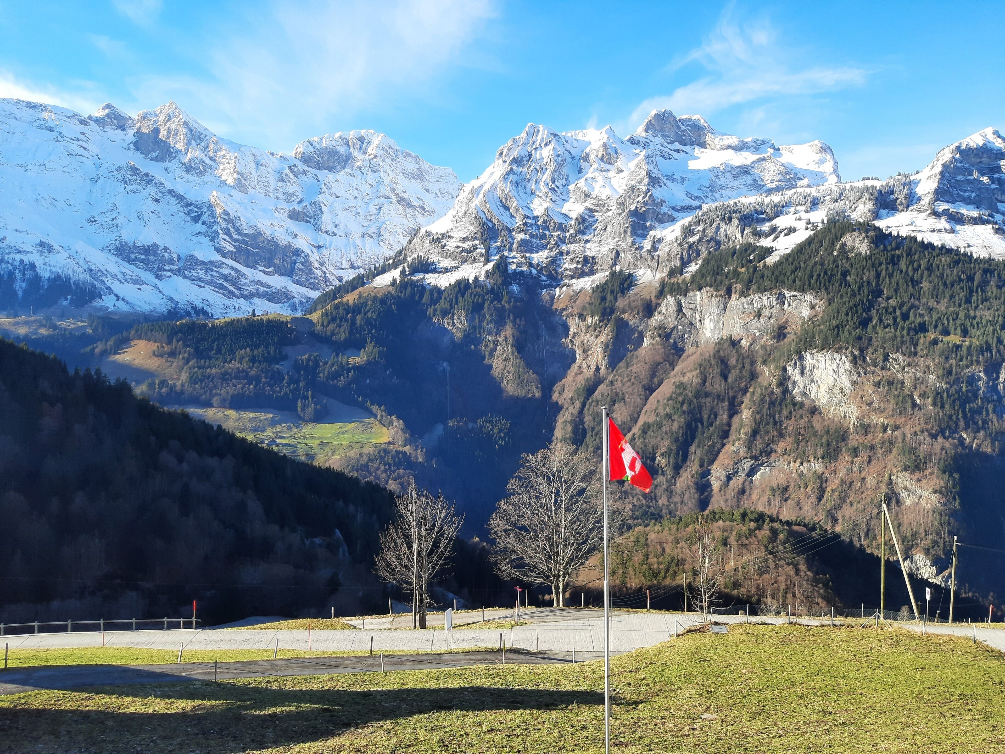 Un paysage montagneux aux sommets enneigés, un mât avec un drapeau, et une zone pavée avec un arbre.