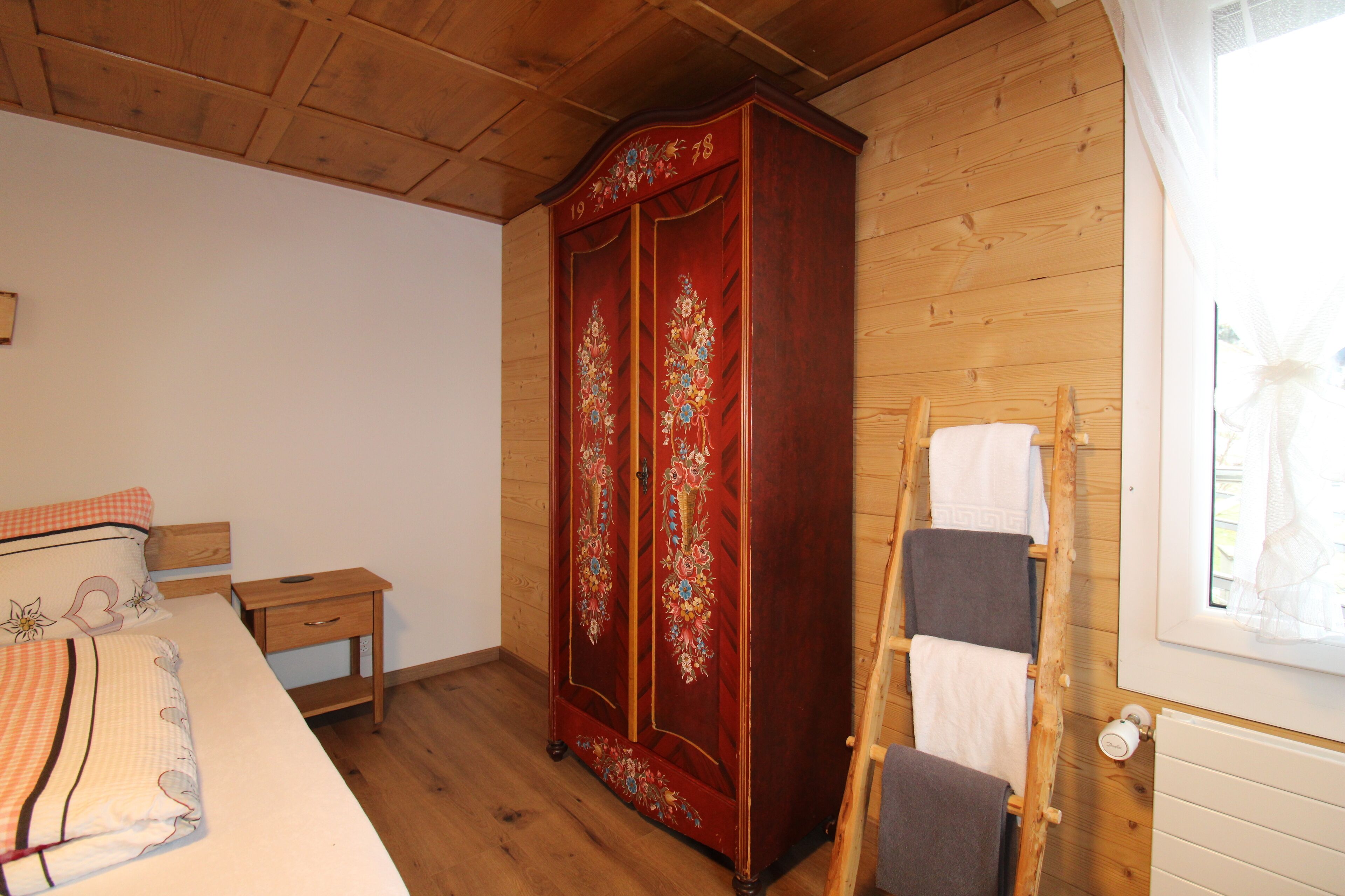 Een slaapkamer met een houten garderobe, een bed met bloemenbeddengoed, een klein nachtkastje en een houten ladder die tegen de muur staat.