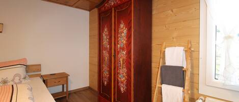A bedroom with a wooden wardrobe, a bed with floral bedding, a small bedside table, and a wooden ladder leaning against the wall.