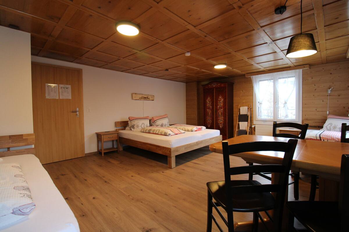 A room with a wooden floor, a bed with pillows, a wooden table with chairs, and a window with curtains.