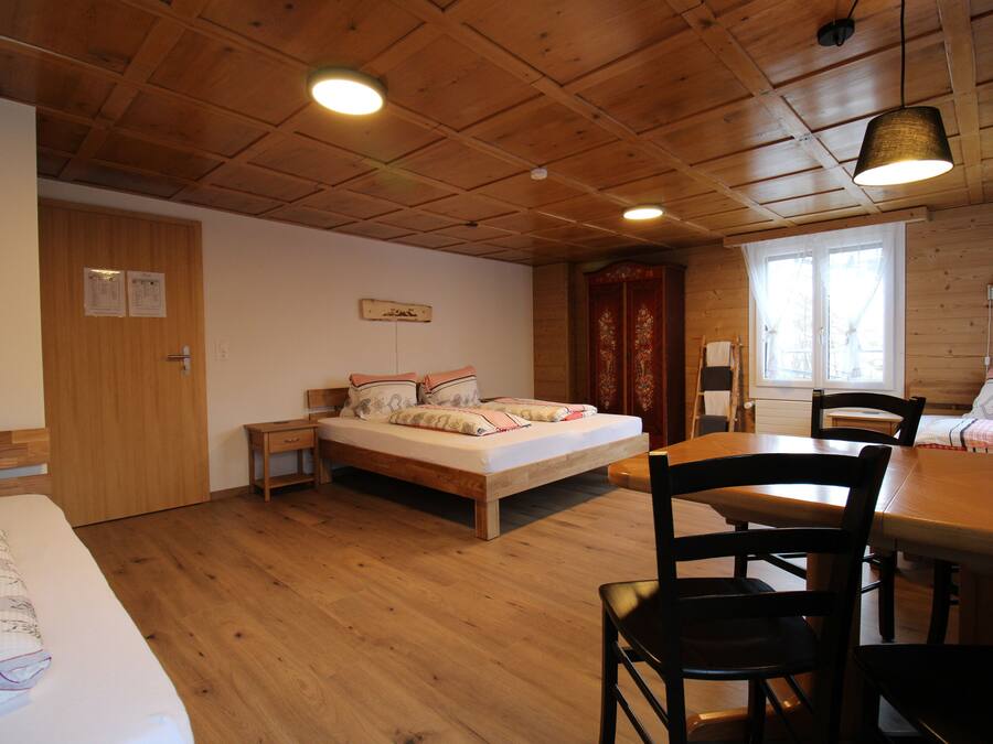 A room with a wooden floor, a bed with pillows, a wooden table with chairs, and a window with curtains.