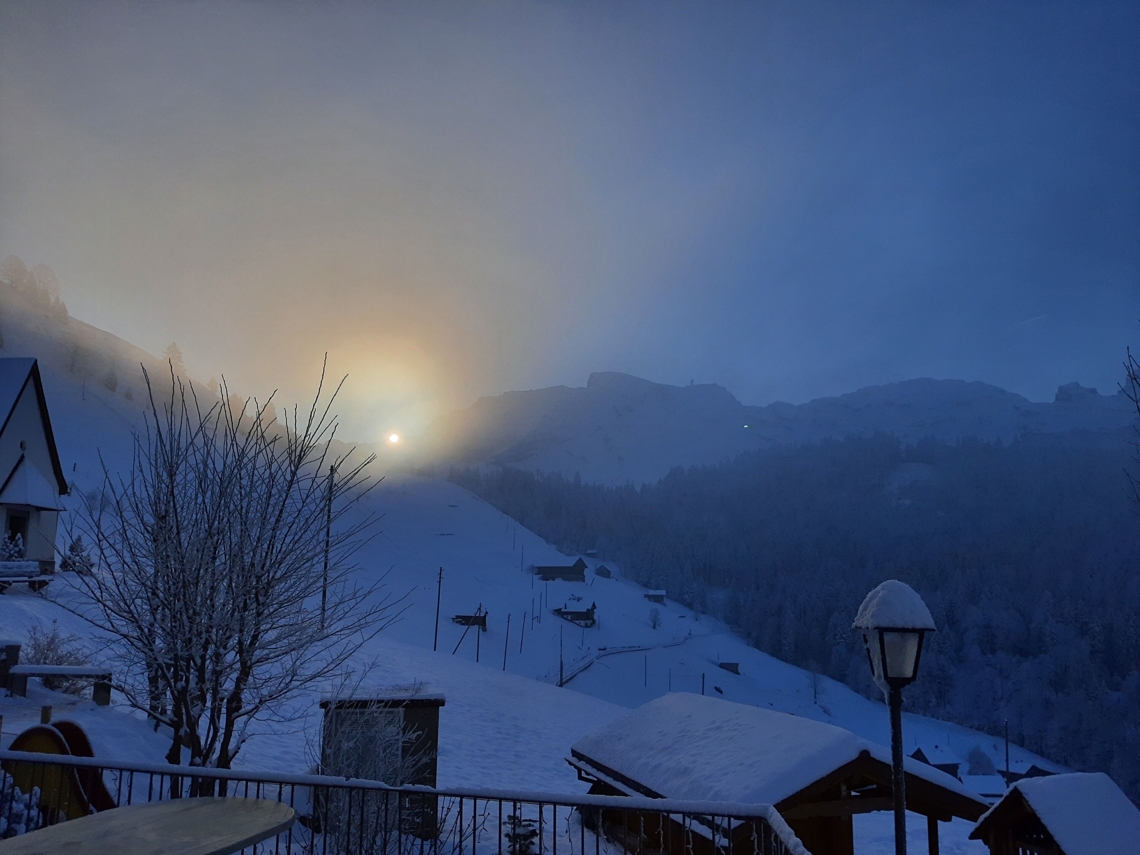 A snowy mountain village at dusk with a visible sun, houses, and a lamppost.