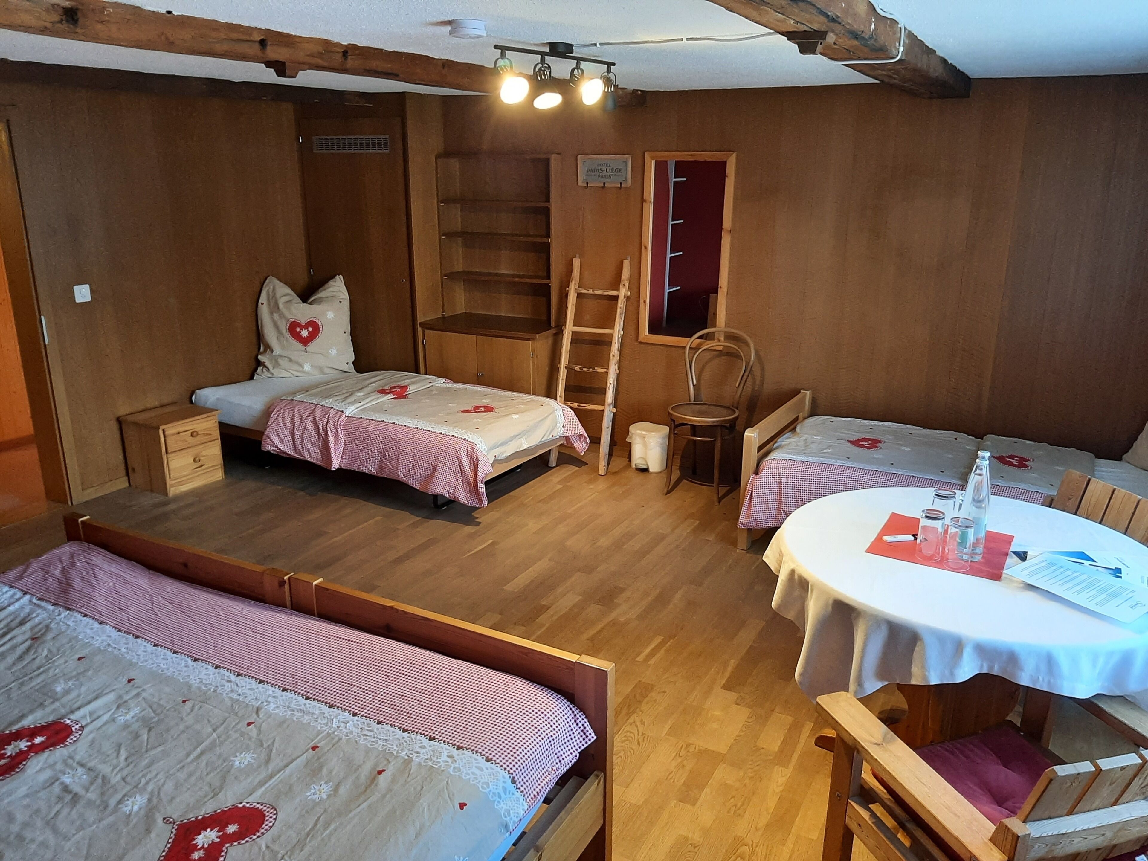 A cozy room with two beds, a small table, and a chair. There is a ladder and a shelf unit.