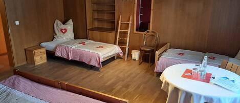 A cozy room with two beds, a small table, and a chair. There is a ladder and a shelf unit.
