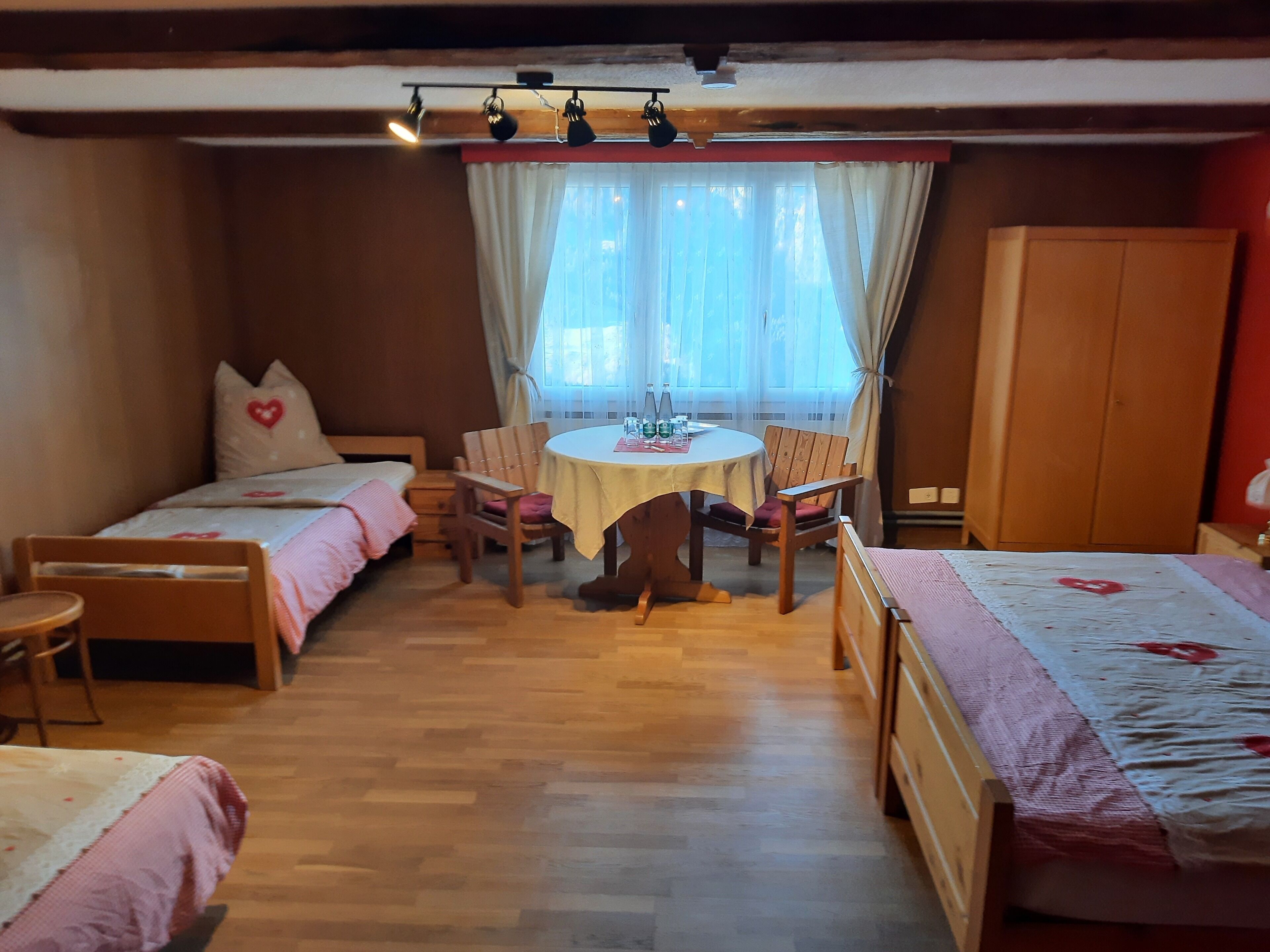 A cozy room with two single beds, a wooden table with chairs, a wardrobe, and a window with curtains.