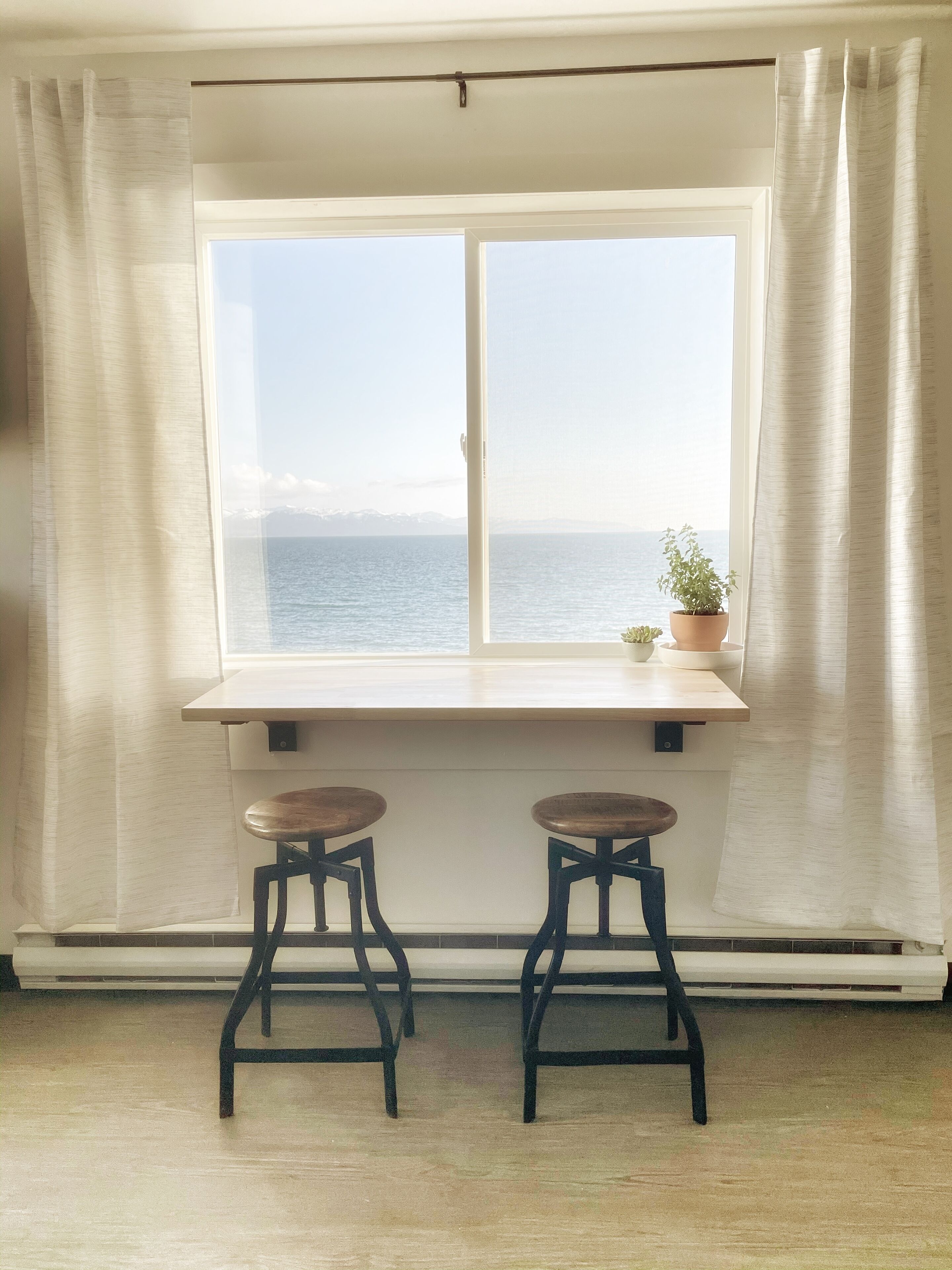 A room with a large window, two wooden stools, and a potted plant on the windowsill.