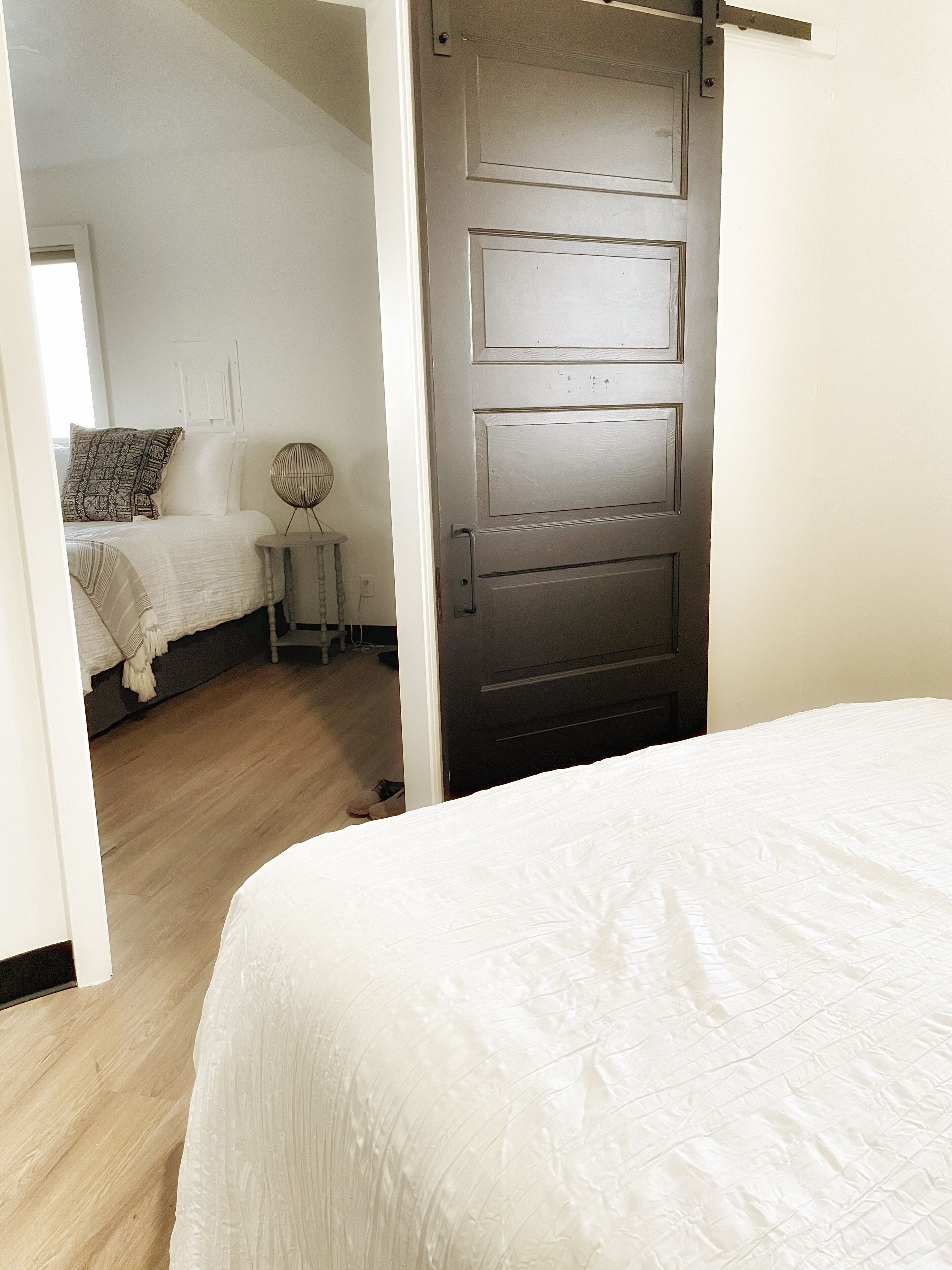 Une chambre à coucher avec un lit, un plancher en bois, une porte et une petite table avec une lampe.