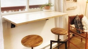 Ein Zimmer mit großem Fenster mit Blick auf einen See und Berge, ein kleiner Tisch mit zwei Hocker und ein Sessel mit Kissen.