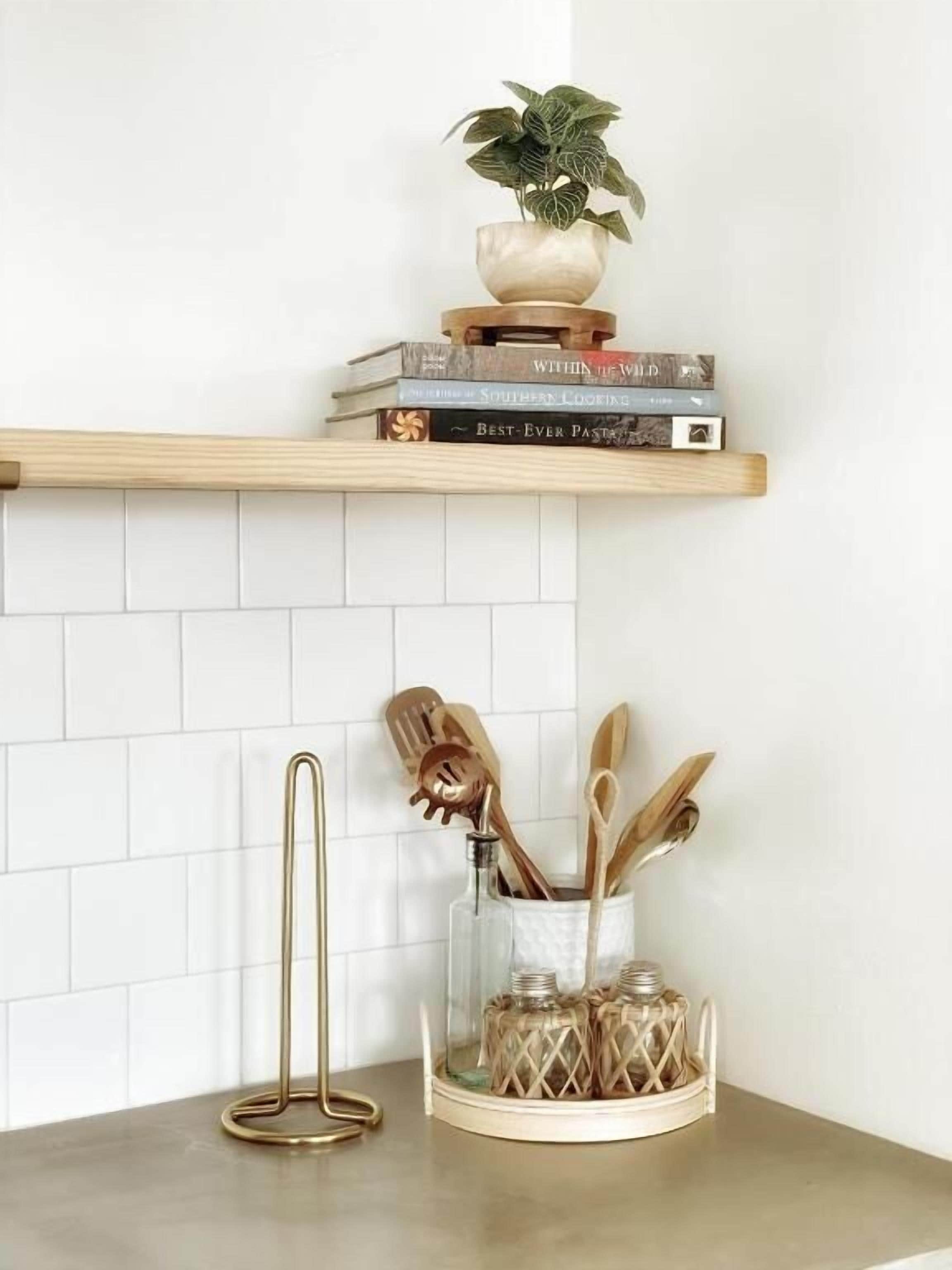 A kitchen counter with a wooden shelf holding books and a potted plant. Below, there is a glass jar with utensils and a gold-colored utensil holder.