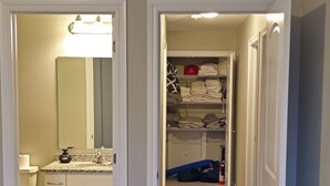 A hallway leading to a bathroom with a toilet, sink, and mirror. There is a closet with shelves and folded clothes.