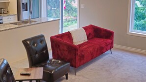 A spacious living area with a red sofa, black leather chairs, and a small wooden table. A kitchen with a refrigerator and a toaster is visible in the background.