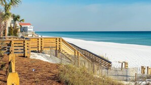 A beach with a wooden boardwalk leading to a white sandy beach, a palm tree, and a building with a red roof in the background. - Beautiful 30A Beach House - short walk to beach! (Santa Rosa Beach)