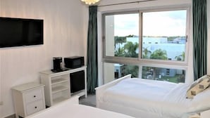 Habitación de hotel con dos camas, un televisor de pantalla plana, un pequeño escritorio con una computadora y una ventana que ofrece vistas a edificios y palmeras.