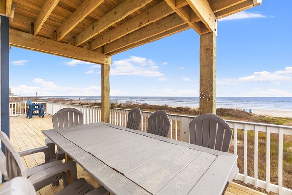 A wooden deck with a table and chairs, overlooking a beach with a clear sky.