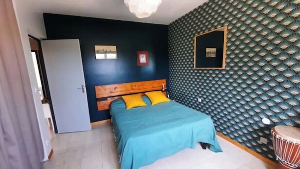 Quarto com cama, cabeceira de madeira, colcha azul e papel de parede estampado.