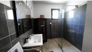 Um banheiro com pia branca, azulejos escuros, armário de madeira e área de chuveiro.