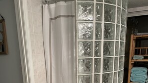 Ein Badezimmer mit einer Duschkabine aus Glas, einem weißen, flauschigen Teppich und einem Holzregal mit gefalteten Handtüchern.