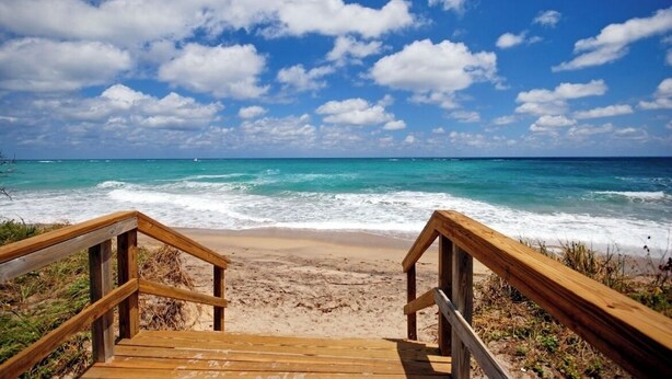 Eine Holztreppe führt zu einem Strand mit blauem Himmel und Meereswellen.