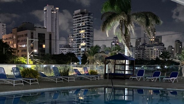 Une piscine avec des chaises longues et un palmier, entourée de grands immeubles la nuit.