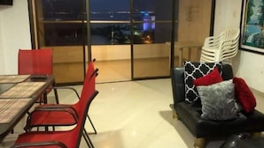 Une pièce comprenant une table à manger, des chaises rouges, un canapé noir agrémenté de coussins à motifs et une grande porte-fenêtre coulissante donnant sur un balcon avec vue sur la ville la nuit.