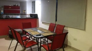Un coin repas avec une table et des chaises, une cuisine à l’arrière-plan et une grande fenêtre avec un store.