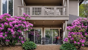 A two-story house with a balcony, a patio with a table and chairs, and large flowering bushes.