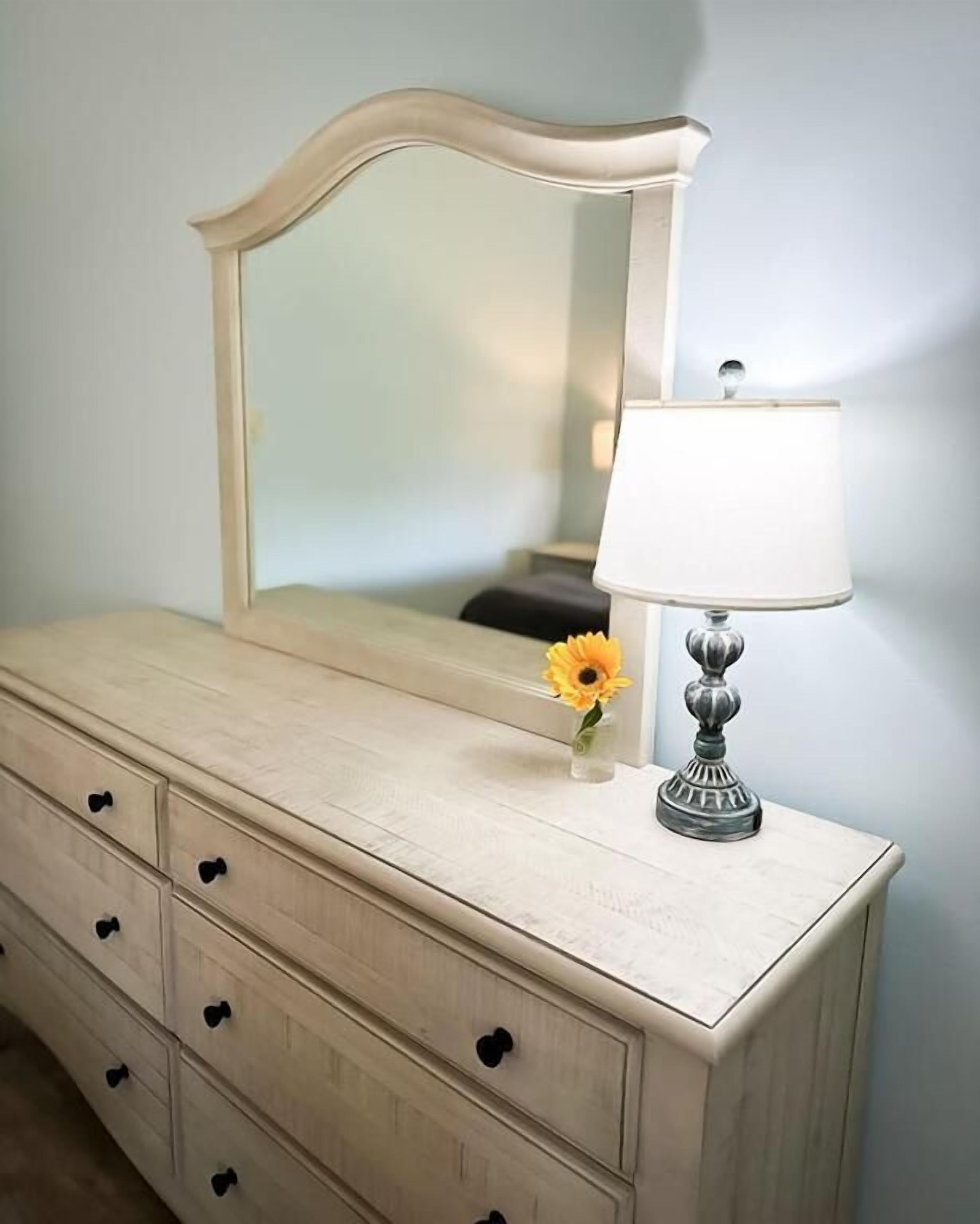 Une commode avec un miroir et une lampe, un vase avec une fleur et un mur avec un interrupteur.
