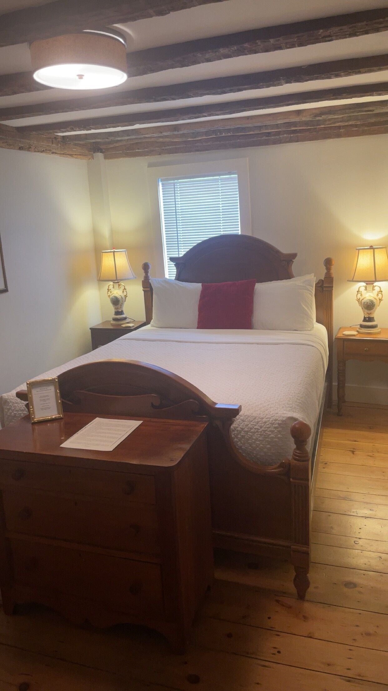 A bedroom with a wooden bed, two bedside tables with lamps, a window with blinds, and a wooden desk with a chair.