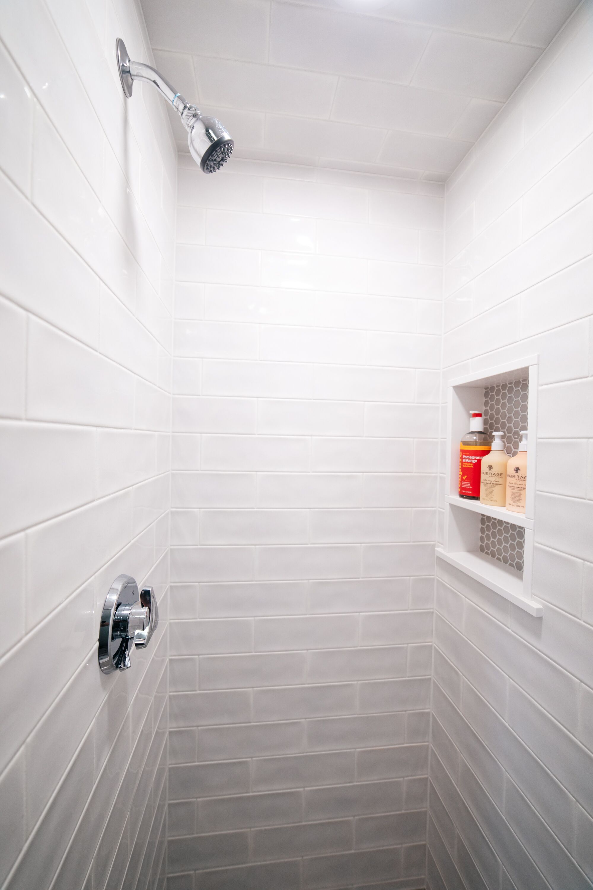 A small, white-tiled shower area with a handheld showerhead and a shelf with toiletries.