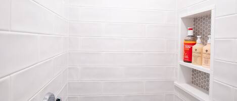 A small, white-tiled shower area with a handheld showerhead and a shelf with toiletries.