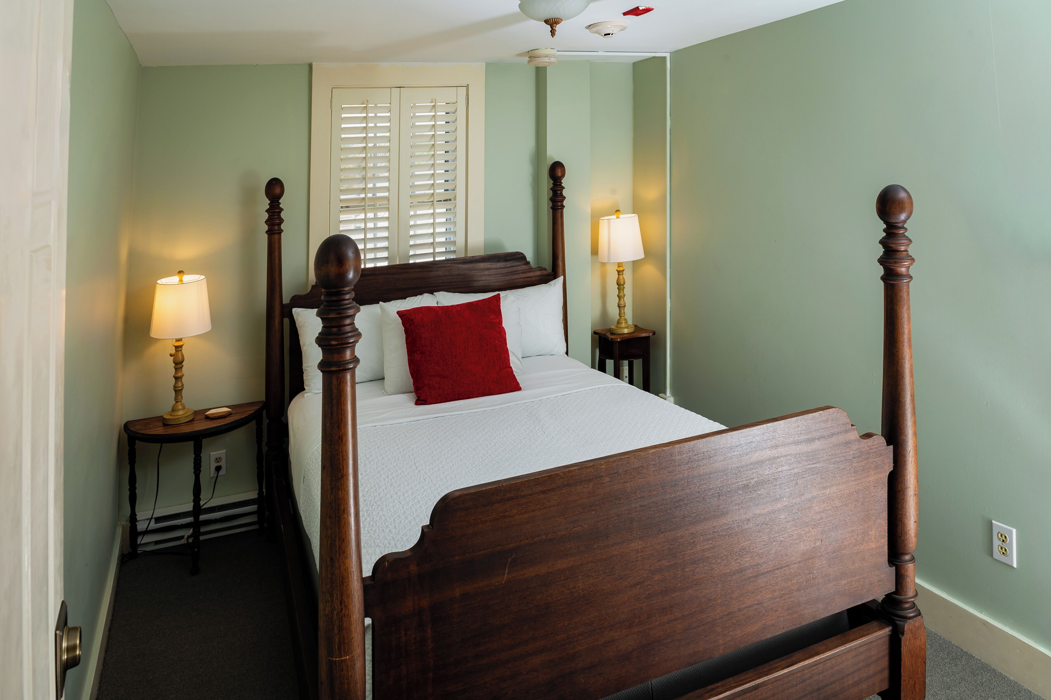 A four-poster bed with a red pillow, two bedside lamps, and a window with blinds.