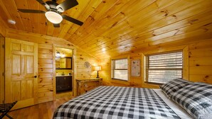 Ein Schlafzimmer in einer Blockhütte mit einem Bett, einem Deckenventilator, Holzwänden, einem Fenster mit Jalousien und einer Tür.