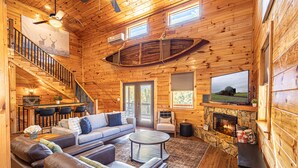 Ein Interieur einer Blockhütte mit einem Wohnzimmer, Kamin, Steinverkleidung des Kamins, Holzbalkendecke und einem großen Fernseher.