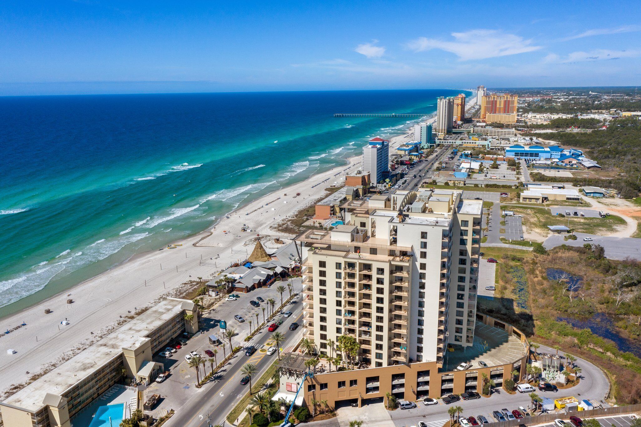 An aerial view of a coastal city with a beachfront hotel, a pier, and high-rise buildings.