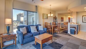 A modern living room with a blue sofa, wooden coffee table, and a dining area with wooden chairs and a small kitchen counter. - Origin 421: 1BR w/ Coastline Views + Steps to Pool! (Panama City Beach)