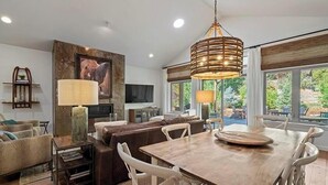 Een moderne woonkamer met een houten eettafel, witte stoelen, een bruine bank, een televisie en een groot raam met uitzicht op groen.