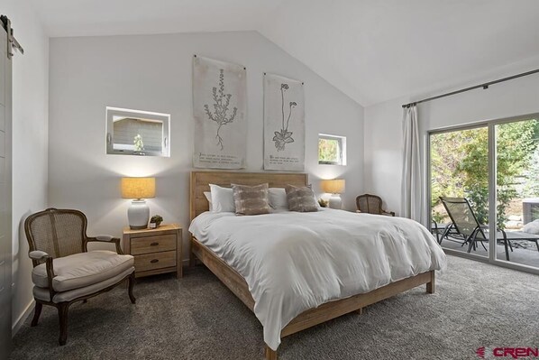 A bedroom with a large bed, two bedside tables, a chair, and a window with a view of greenery. - Hot Tub, Fireplace, 15 Minutes from Skiing! (Durango)