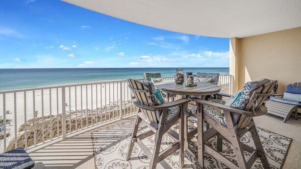 En balkon med udsigt over stranden og havet, møbleret med et bord og stole, samt en gulvtæppe med et mønster.